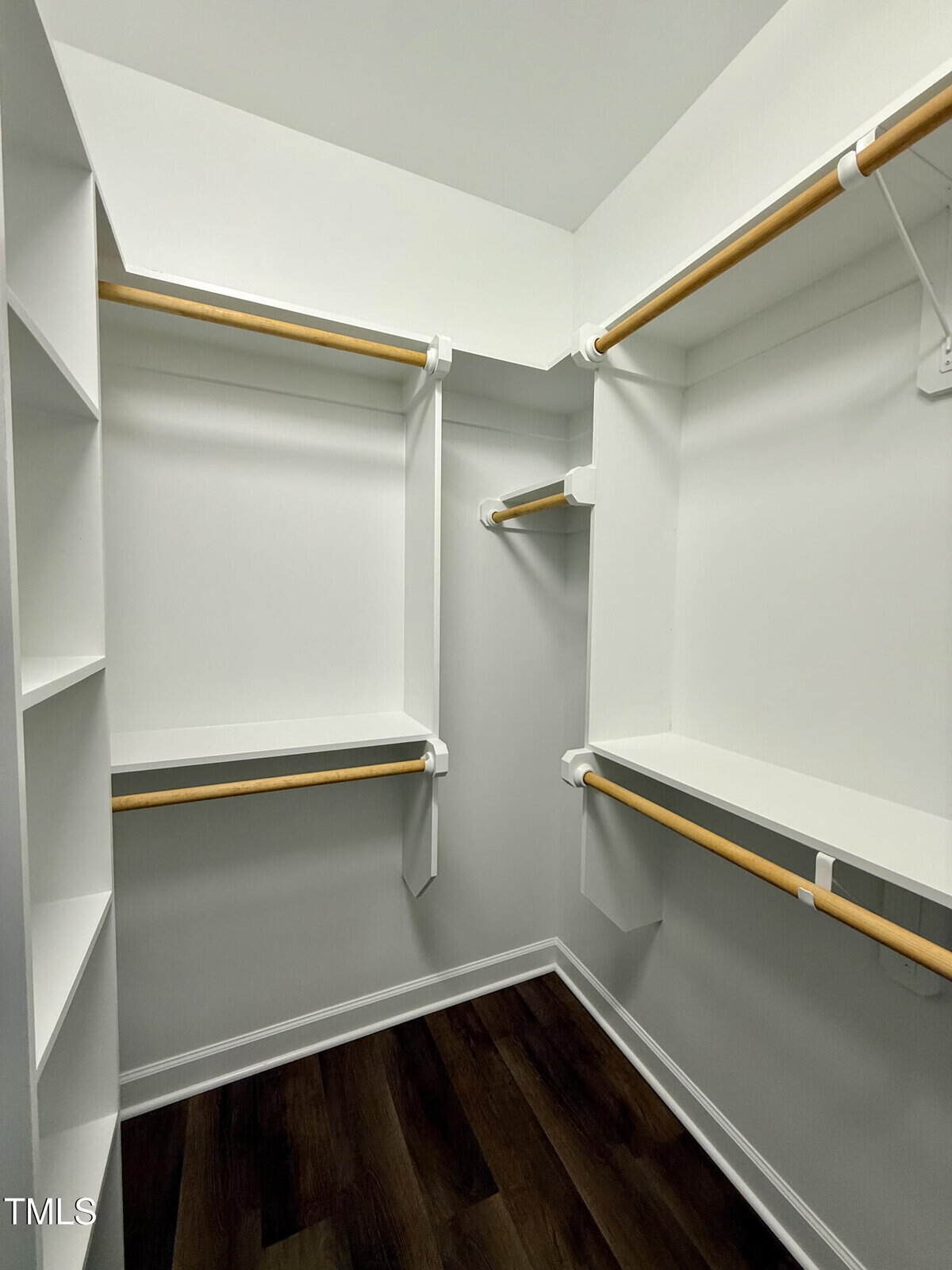 A walk-in closet with white shelves and hanging rods on three walls, featuring dark wood flooring and white walls. The closet includes multiple storage sections and open shelving.