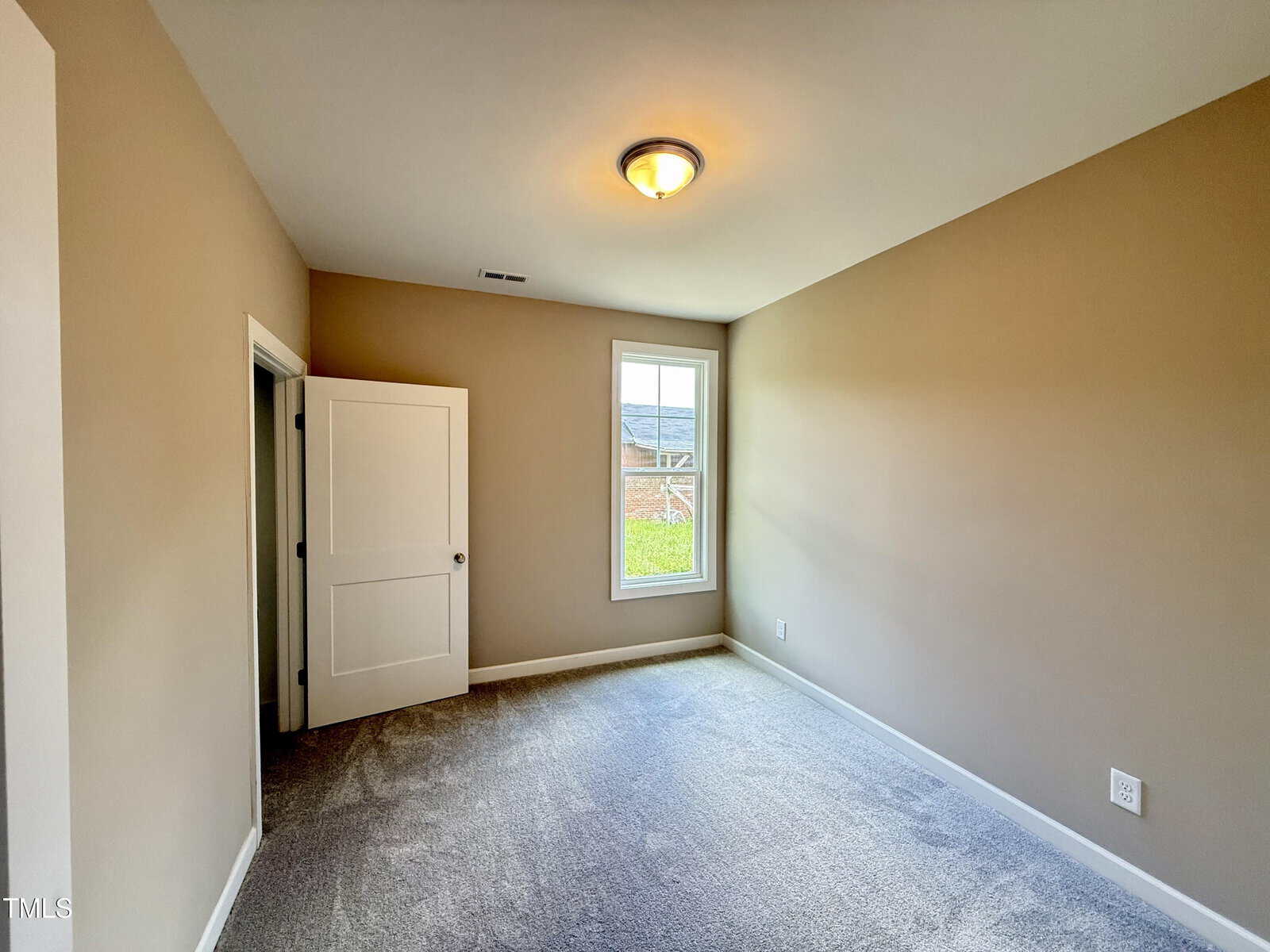 A small, empty room with beige walls, gray carpet, a ceiling light, a closed white closet door, and a window letting in natural light.