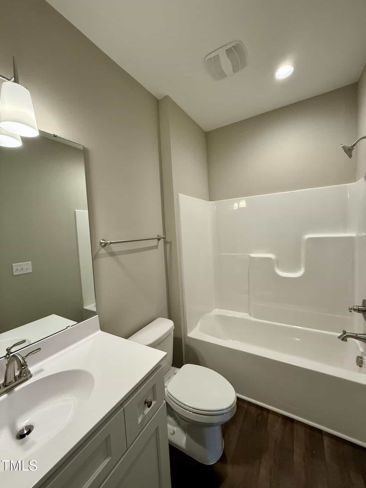A small bathroom with a white vanity and sink, a mirror above, a toilet, and a shower-tub combo with a built-in shelf. The walls are beige, and the floor is dark wood or wood-look material.