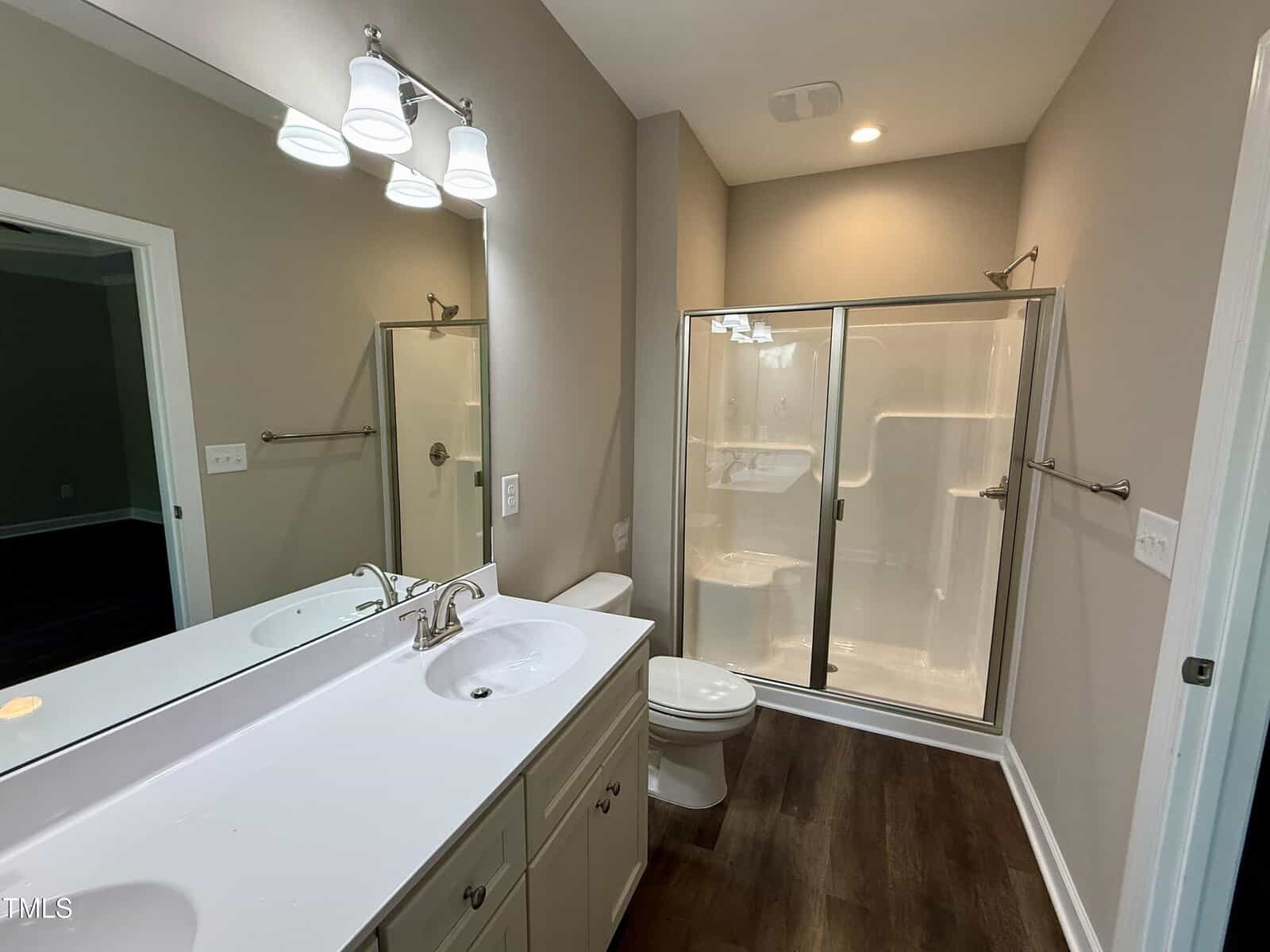 A bathroom with a white vanity and sink, large mirror, toilet, and a glass-enclosed shower. The walls are beige, the floor is dark wood, and the space is well-lit with overhead lights.