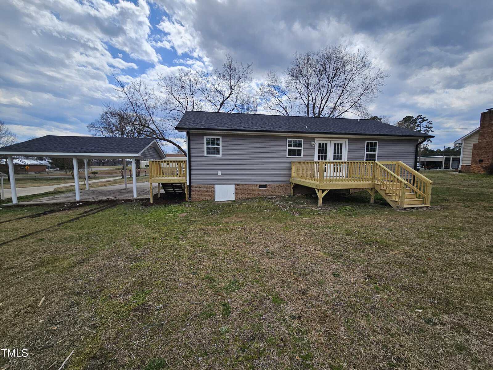 A single-story gray house with a black roof and wooden deck is situated on a grassy lawn. There is a covered carport to the left, and a chimney is visible on the right. The sky is partly cloudy.