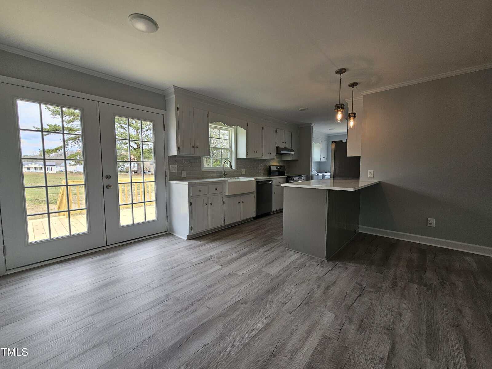 A modern kitchen features light gray hardwood floors, white cabinetry, and a center island with a counter. Pendant lights hang above the island. Large glass doors on the left lead to an outdoor area with a tree visible outside.