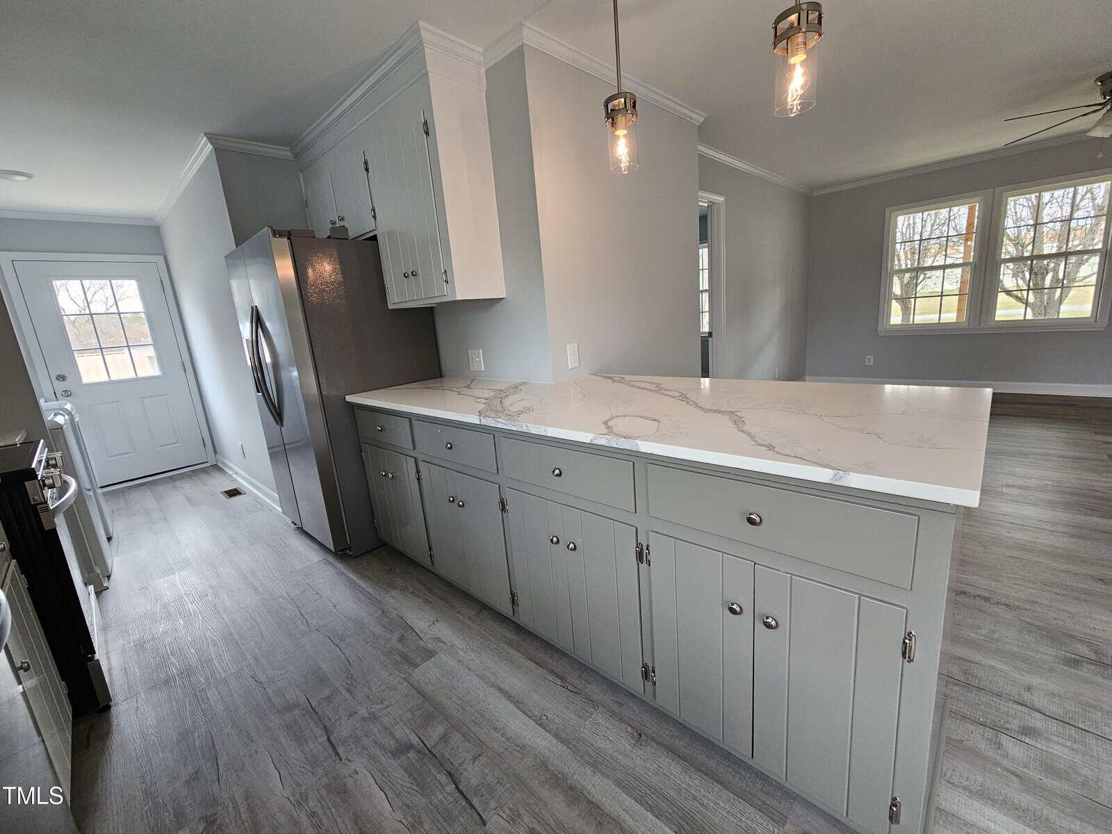A modern kitchen with light gray cabinets and a marble countertop island. Stainless steel refrigerator is to the left, and pendant lights hang above the island. Large windows and a door provide natural light from the back wall. Floors are light wood.