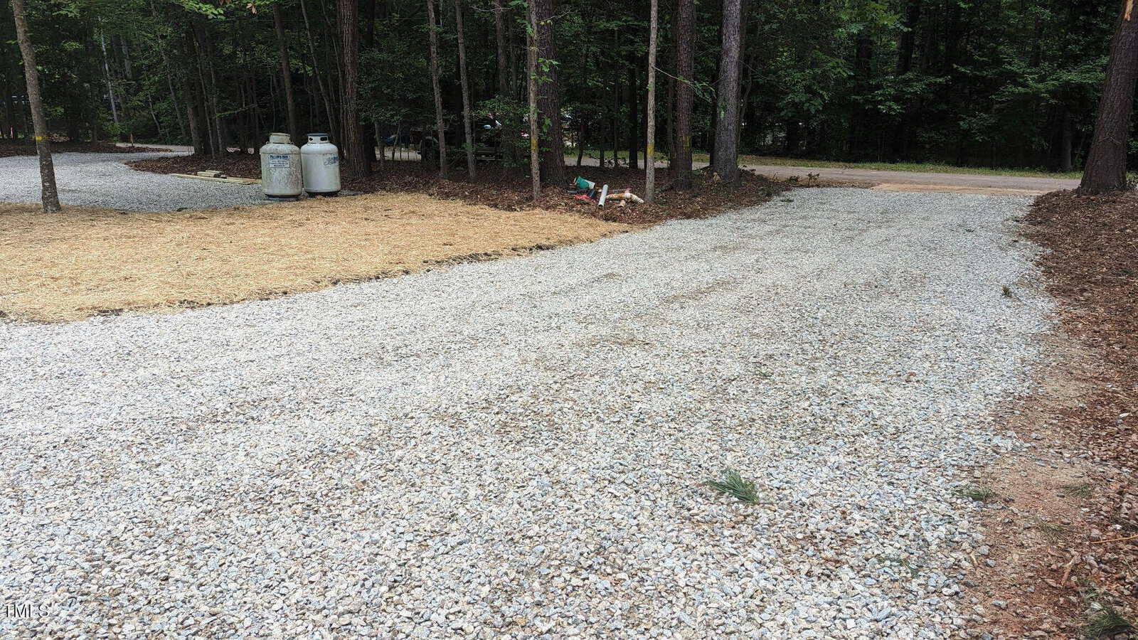 A gravel driveway curves through a wooded area with propane tanks and utility connections on the left. Trees line the driveway, and there is a road in the background.