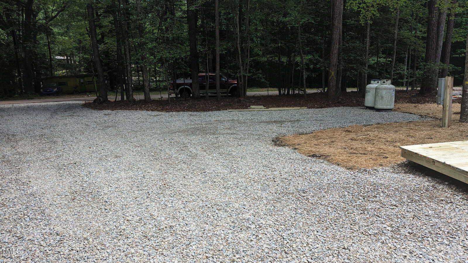 A gravel driveway leads to a wooded area with trees at 110 112 Chama Drive in Louisburg. Two large propane tanks sit on the right near a wooden platform, while a black truck is parked near the trees in the background.