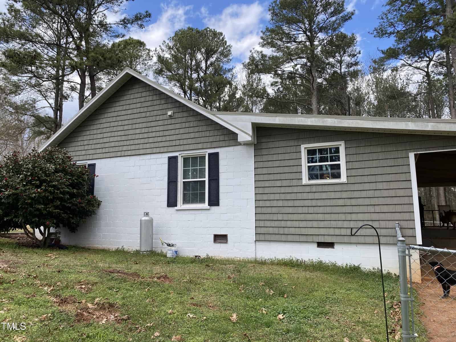 A single-story house with gray shingle siding and white painted lower walls. There are two windows, black shutters, a propane tank, and a fenced yard with grass and trees in the background.