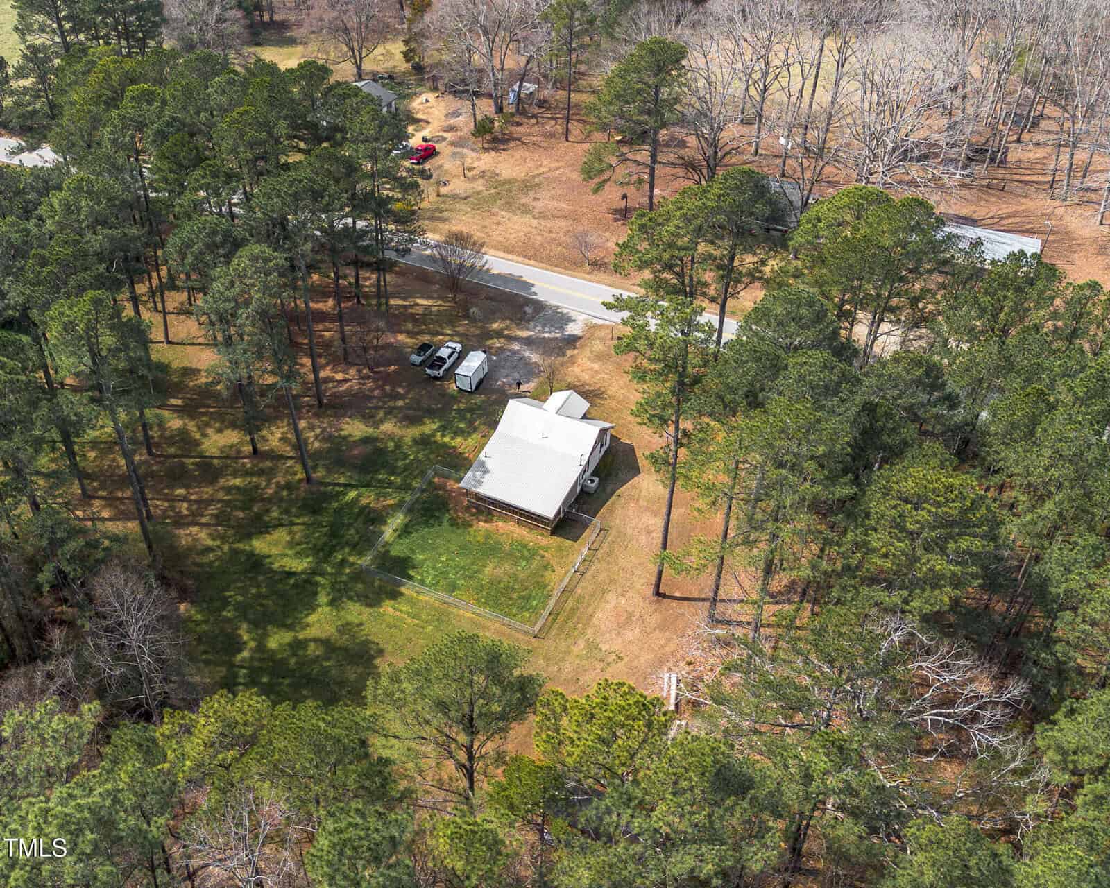 Aerial view of a white house with a fenced yard, surrounded by tall trees. Two vehicles are parked next to the house, and a paved road runs nearby through a wooded area.