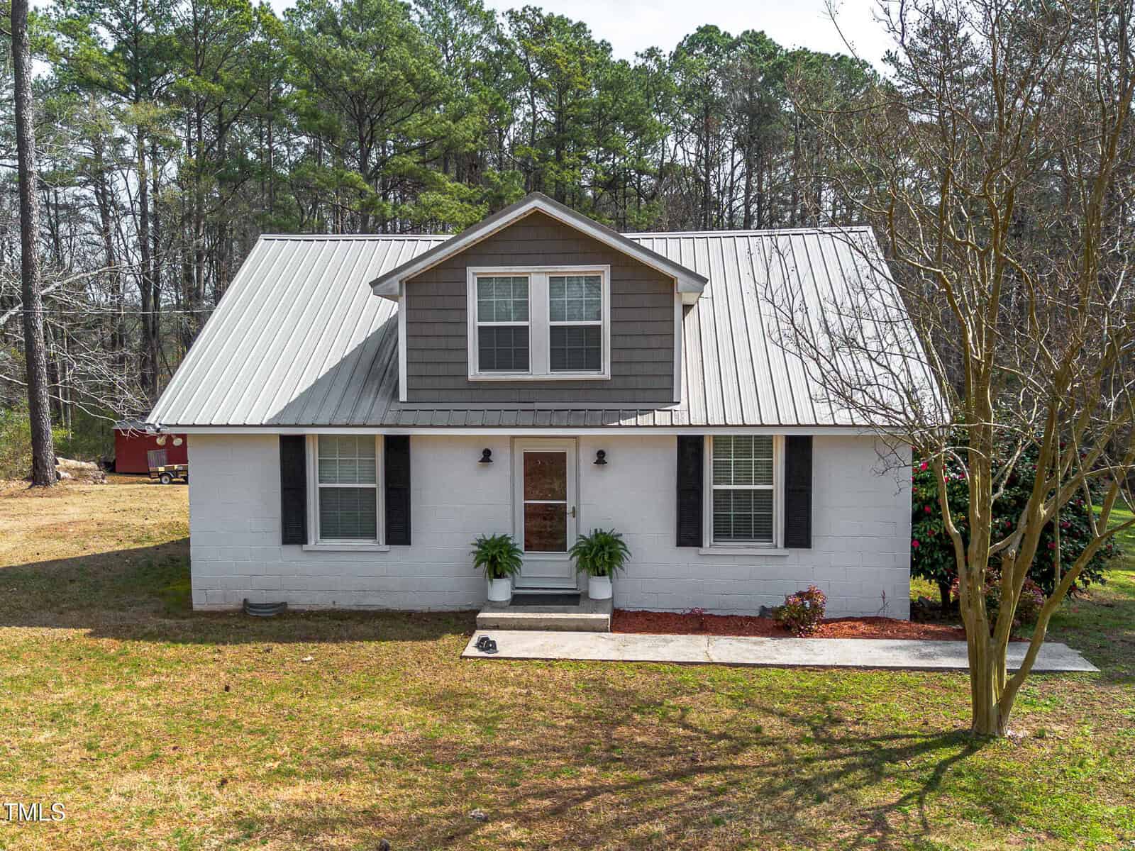 A white, one-and-a-half-story house with a gray metal roof and dormer window, two front windows with black shutters, two small potted plants by the entrance, and a leafless tree in the front yard. Pine trees are in the background.