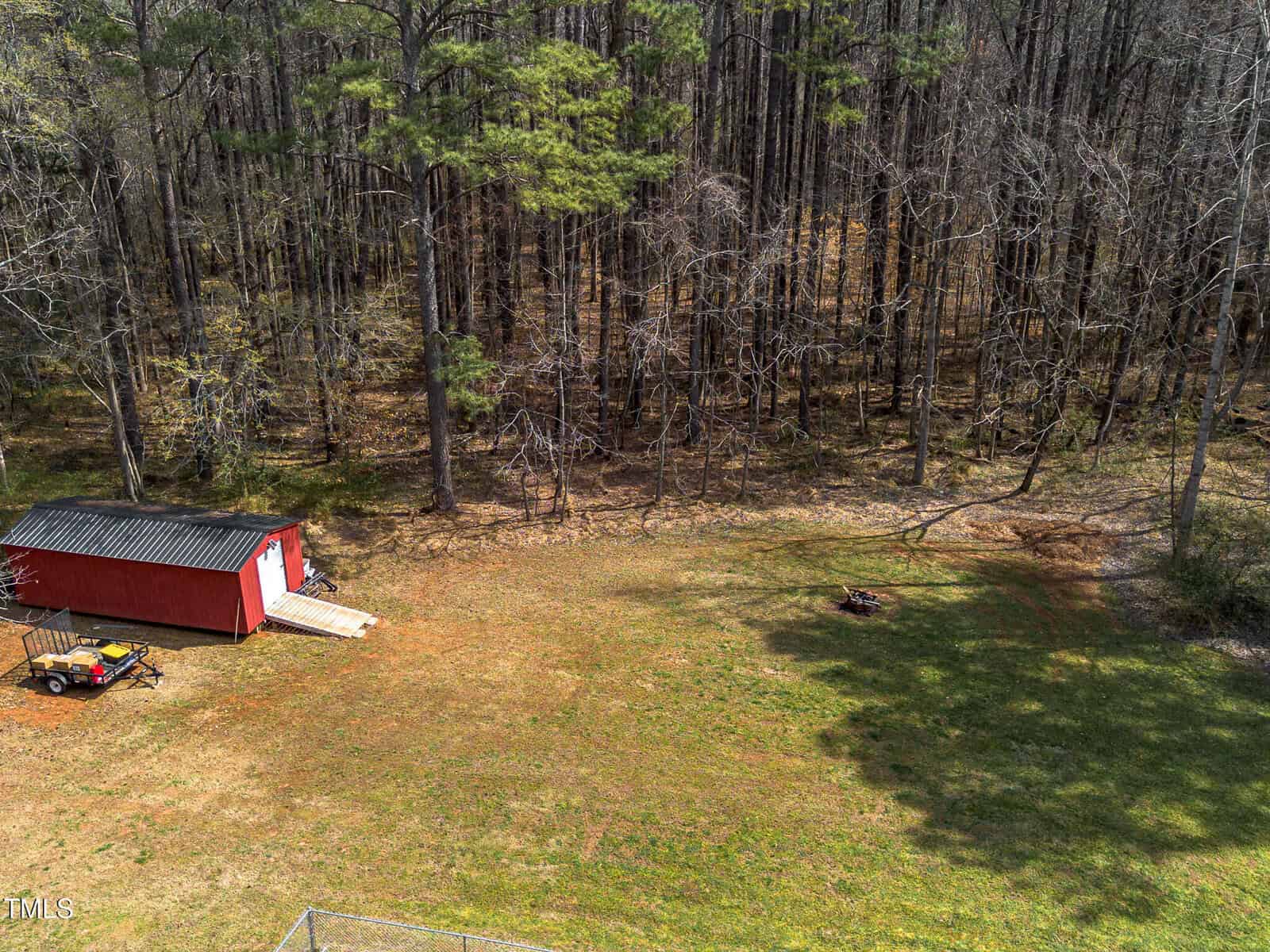 A red shed with a black roof sits on a grassy area by the edge of a forest. A ramp leads to the shed door. A small trailer is parked nearby, and there is a firepit on the grass. Tall trees fill the background.