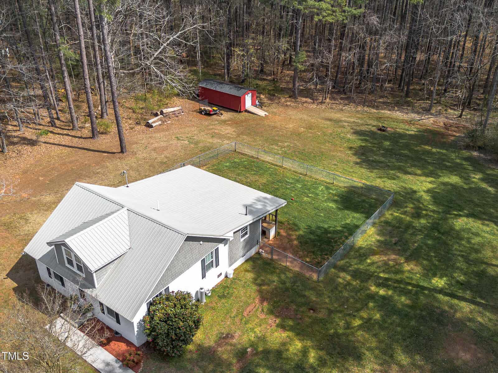 Aerial view of a white house with a metal roof, a fenced backyard with green grass, and a red shed near a wooded area. The property is surrounded by trees and open lawn.