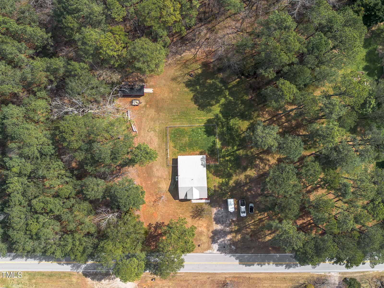 Aerial view of a house with a light roof surrounded by trees, a small outbuilding, two parked vehicles, a visible road at the bottom, and a rectangular lawn area behind the house.