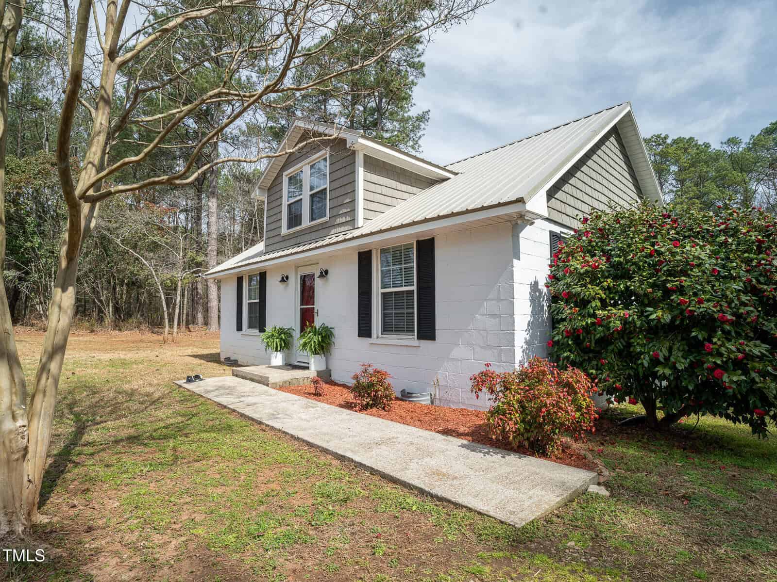 A small two-story house with gray siding and white brick exterior, a metal roof, red front door, shrubs, and a concrete walkway. Trees and grass yard surround the home under a partly cloudy sky.