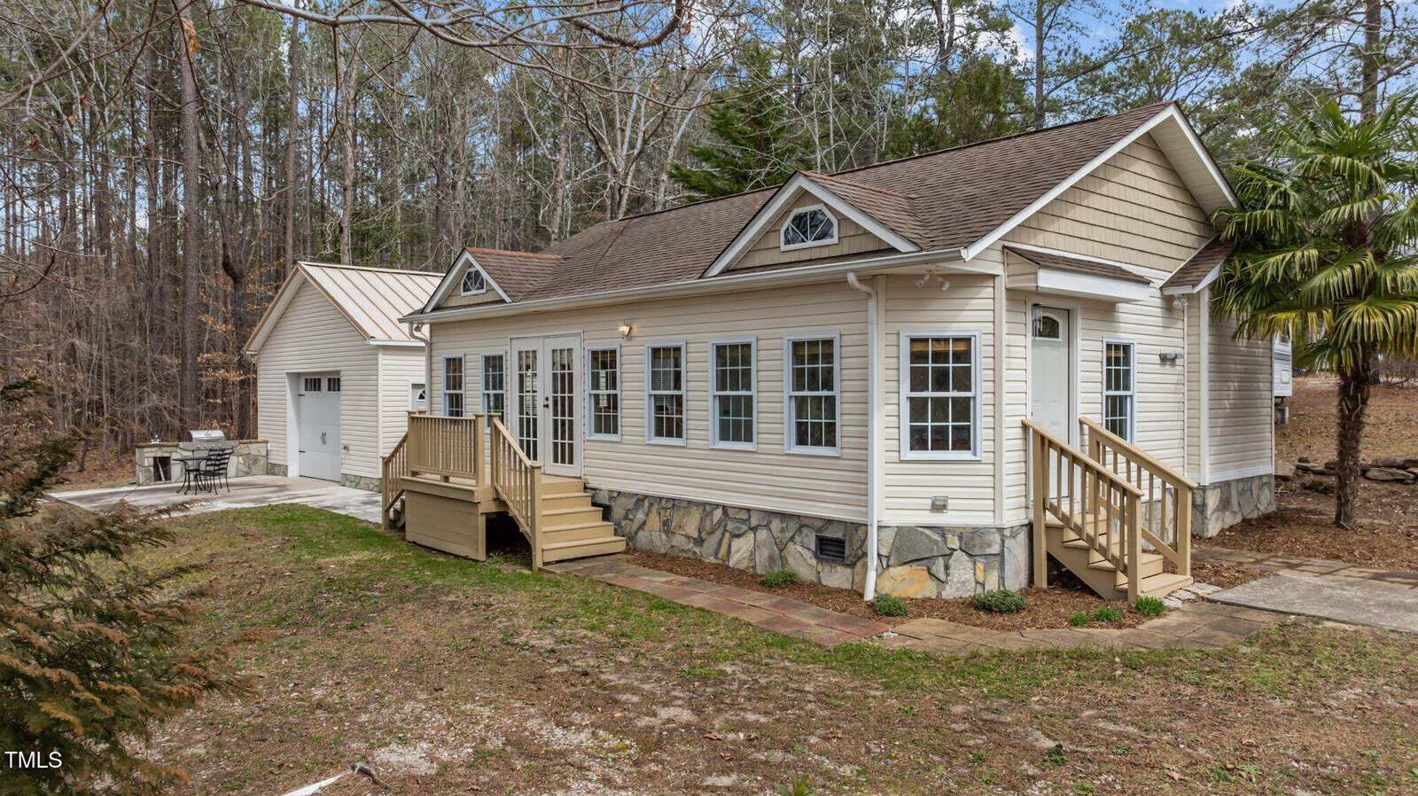 Single-story beige house with stone foundation, two small decks, and multiple windows. The house sits on a grassy lot with surrounding trees, and a detached garage is visible in the background.