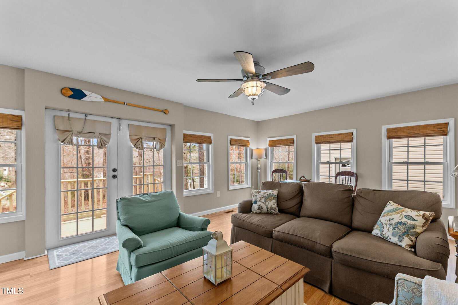 A living room with a brown sofa, green armchair, and wooden coffee table. There are several windows with bamboo shades, a ceiling fan, and double doors leading to a deck. An oar is mounted above the doors.