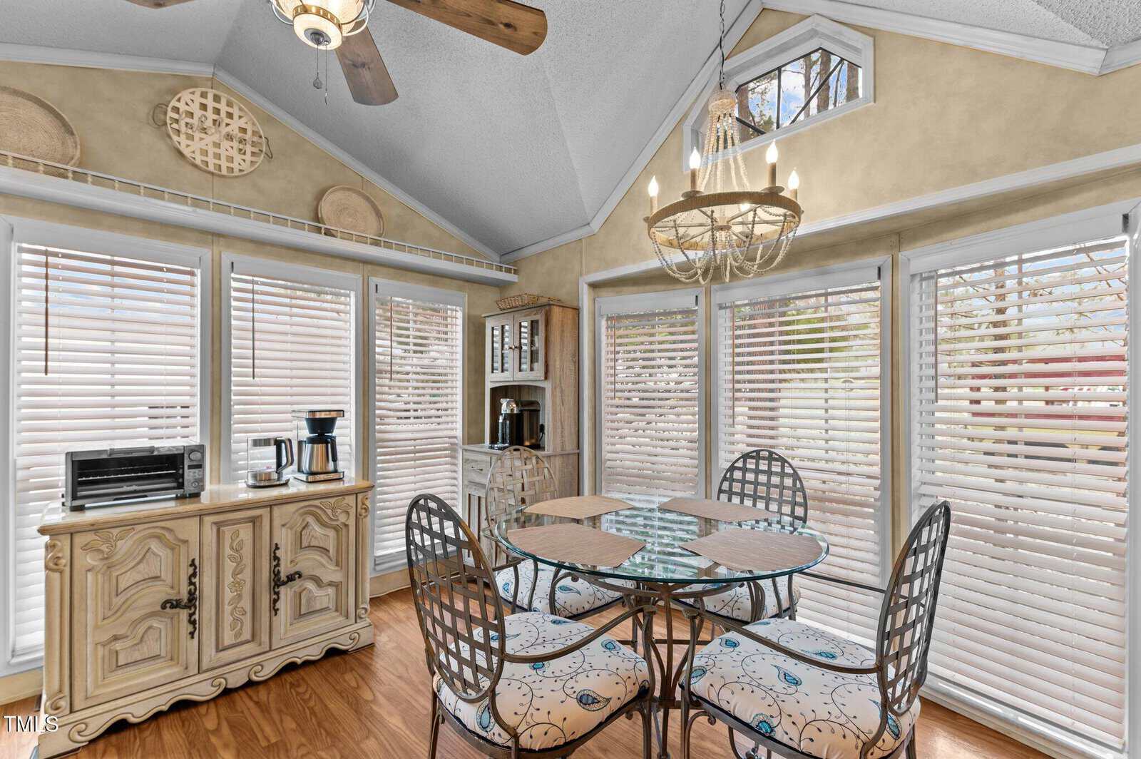 Breakfast nook with a round glass table, four metal chairs, and a chandelier. A sideboard with a microwave and coffee maker sits against windows with white blinds. Wood floors and neutral walls are visible.