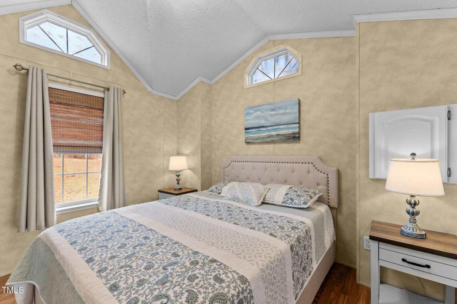 A bedroom with beige walls, a large bed with a patterned comforter, two nightstands with lamps, windows with curtains, and a seascape painting above the bed. Hardwood floor and vaulted ceiling are visible.