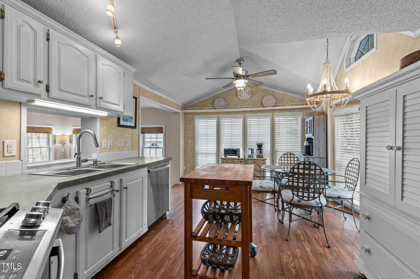 A kitchen with white cabinets, a double sink, and an island leads into a dining area with a glass table and metal chairs. There are large windows with blinds, a ceiling fan, a chandelier, and wood flooring throughout.