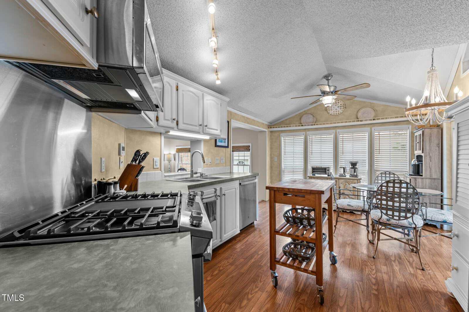 A kitchen with white cabinets, stainless steel appliances, and a gas stove, with a wooden rolling island in the center. A small dining area with a table and metal chairs is by large windows in the background.