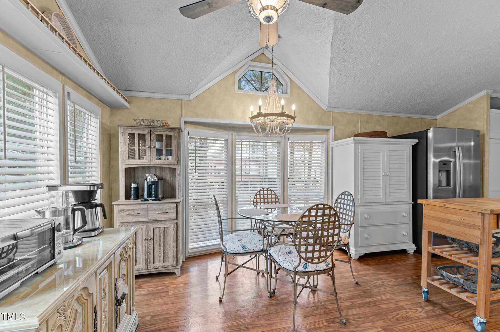 Bright kitchen with wooden floors, metal dining table and chairs, coffee station, refrigerator, island cart, chandelier, and large windows with white blinds. Ceiling fan and high ceiling with triangular window are visible.