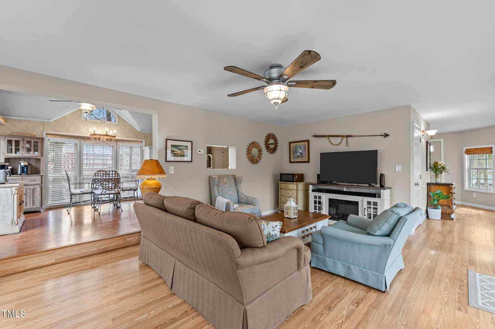 A living room with wood floors, a ceiling fan, two couches, an armchair, a TV on a white stand, and decor including mirrors and framed art; a kitchen and dining area are visible in the background.