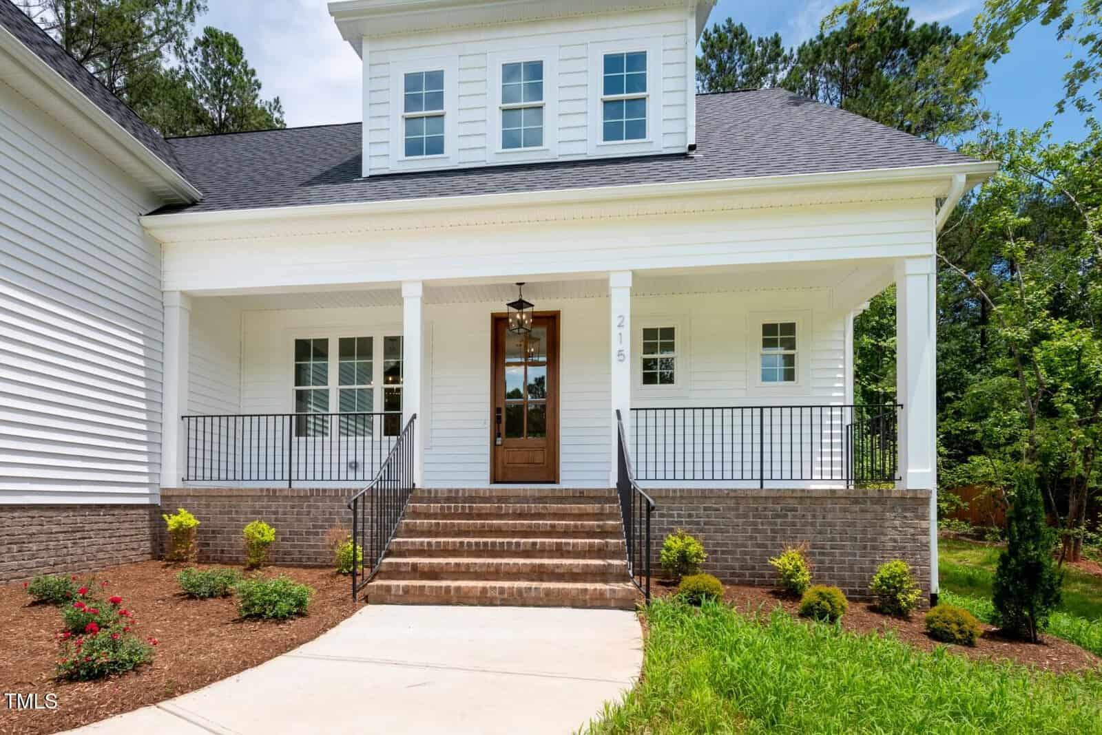 A white house with a covered front porch, wooden steps, black railings, and landscaped bushes. The porch has two white columns, two windows, and a central door with a hanging light fixture above.