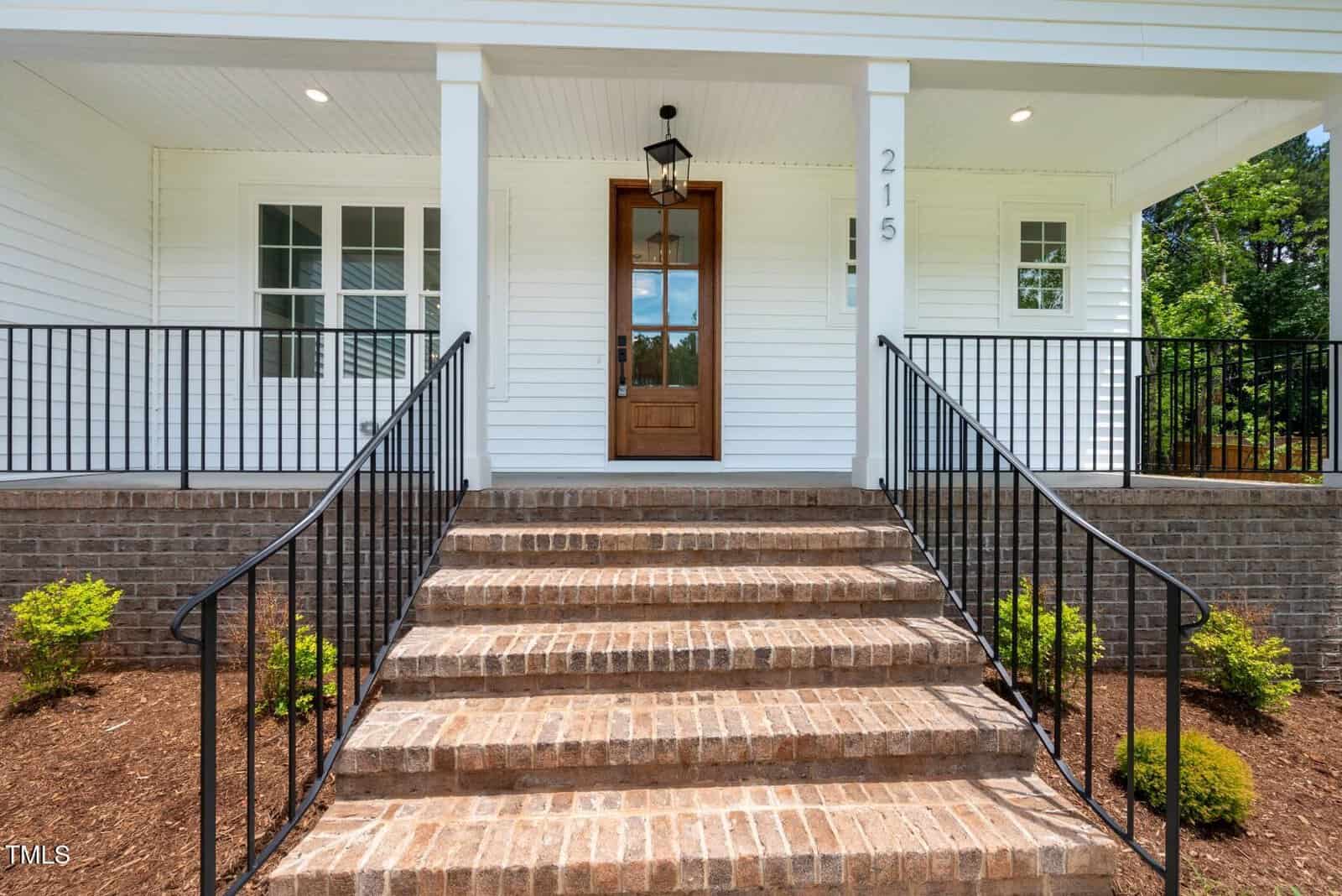 Front porch of a house with brick steps, black railings on both sides, a wooden front door with glass panels, white siding, windows on the left, and house number 215 on a column. Shrubs are planted along the steps.