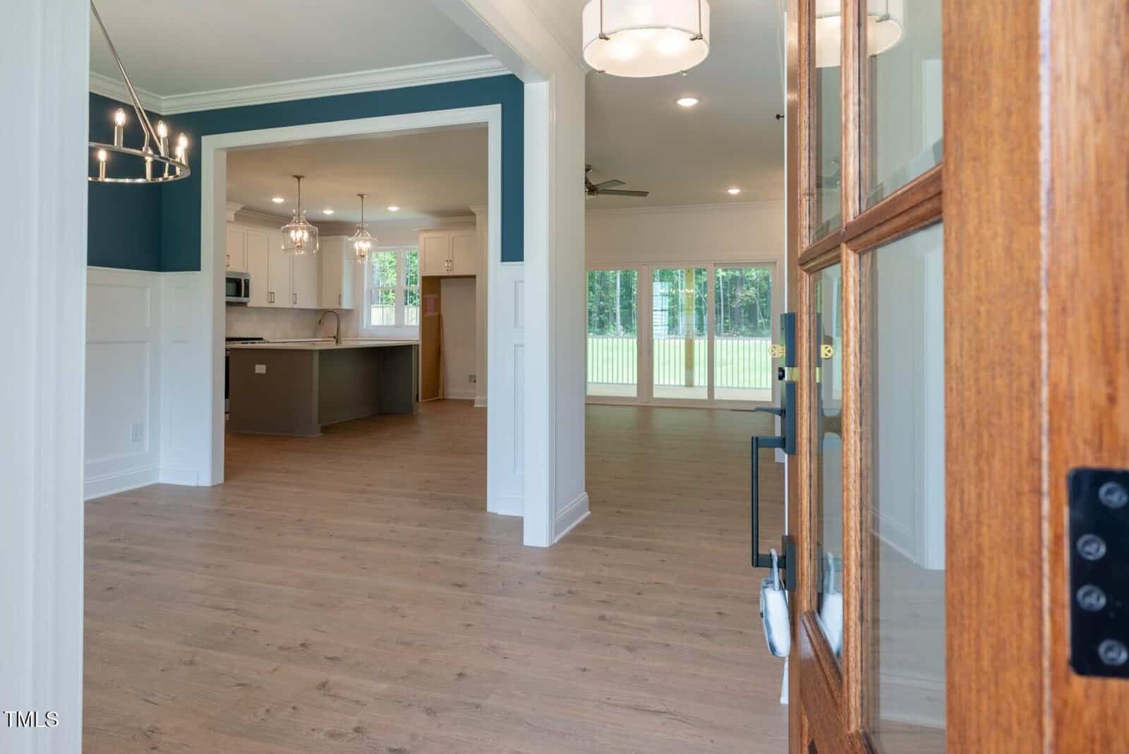 View from an open wooden door into a modern home with light wood floors, white walls, teal accent, two chandeliers, and an open kitchen and living area with large windows showing a green yard.