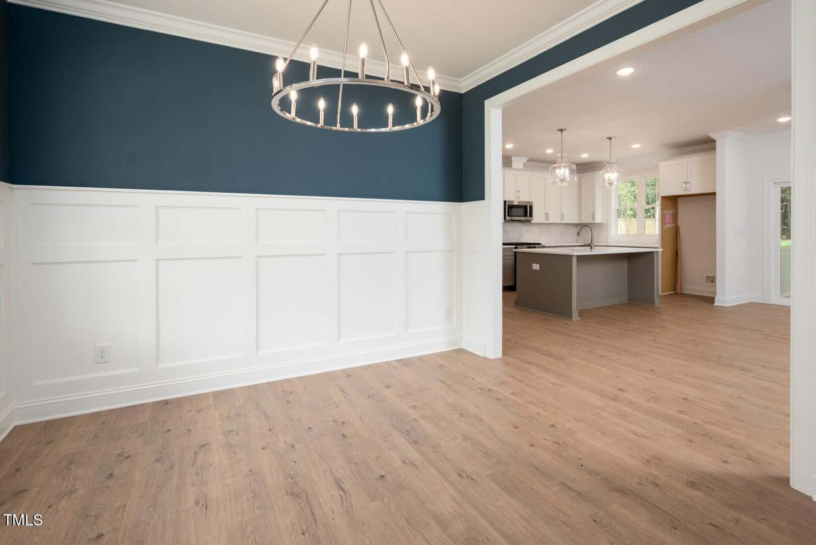 A dining area with wood flooring and white wainscoting, featuring a modern chandelier. The space opens to a kitchen with white cabinets, an island, stainless steel appliances, and large windows.