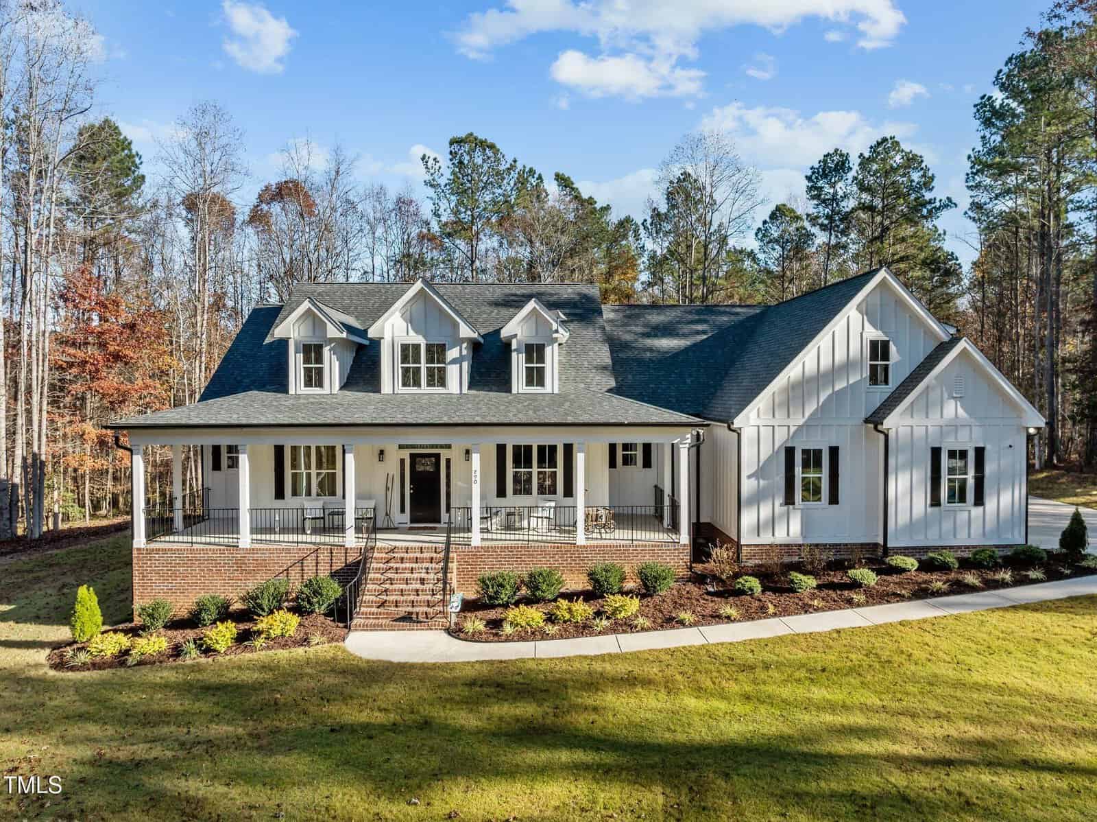 A two-story white house with black roof, dormer windows, and a large front porch with columns and railings, set on a spacious lawn with landscaped shrubs, trees, and a curved walkway.