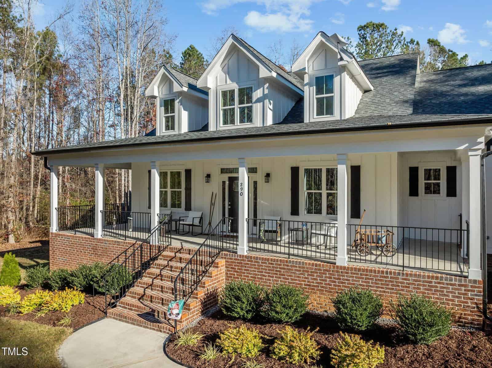 A two-story white house with a large covered front porch, black railings, brick steps and foundation, dormer windows, and neatly landscaped bushes, surrounded by trees under a blue sky.