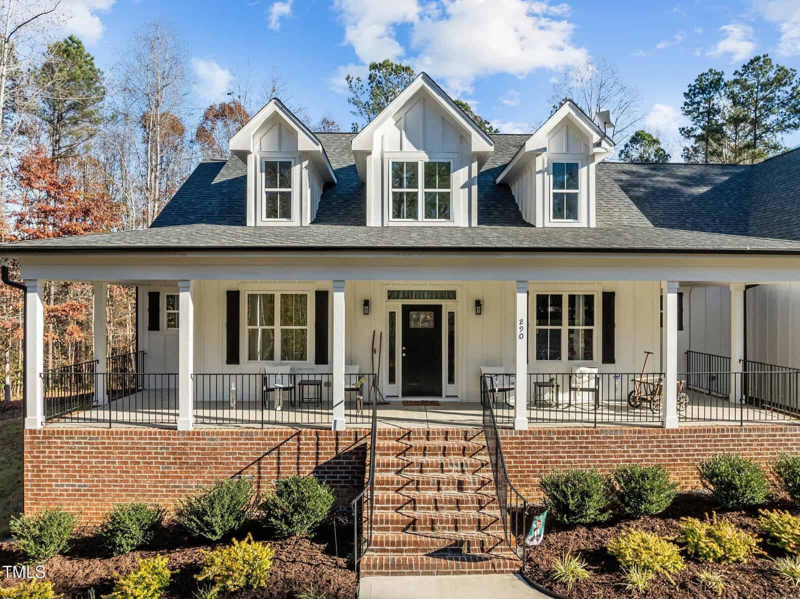 A modern farmhouse with white siding, brick foundation, dormer windows, and a large covered front porch with black railings. There are steps leading up to the porch and landscaped bushes in front.