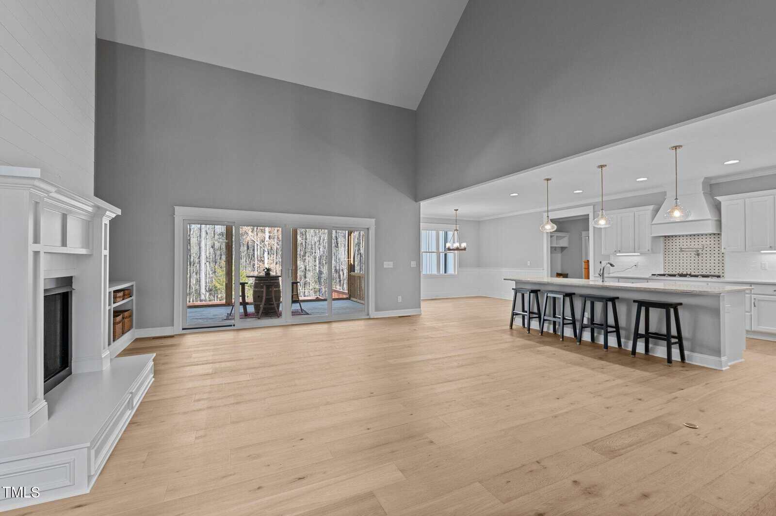 Spacious open-concept living area with light wood floors, a white fireplace on the left, large sliding glass doors at the back, and a modern kitchen with an island, pendant lights, and bar stools on the right.