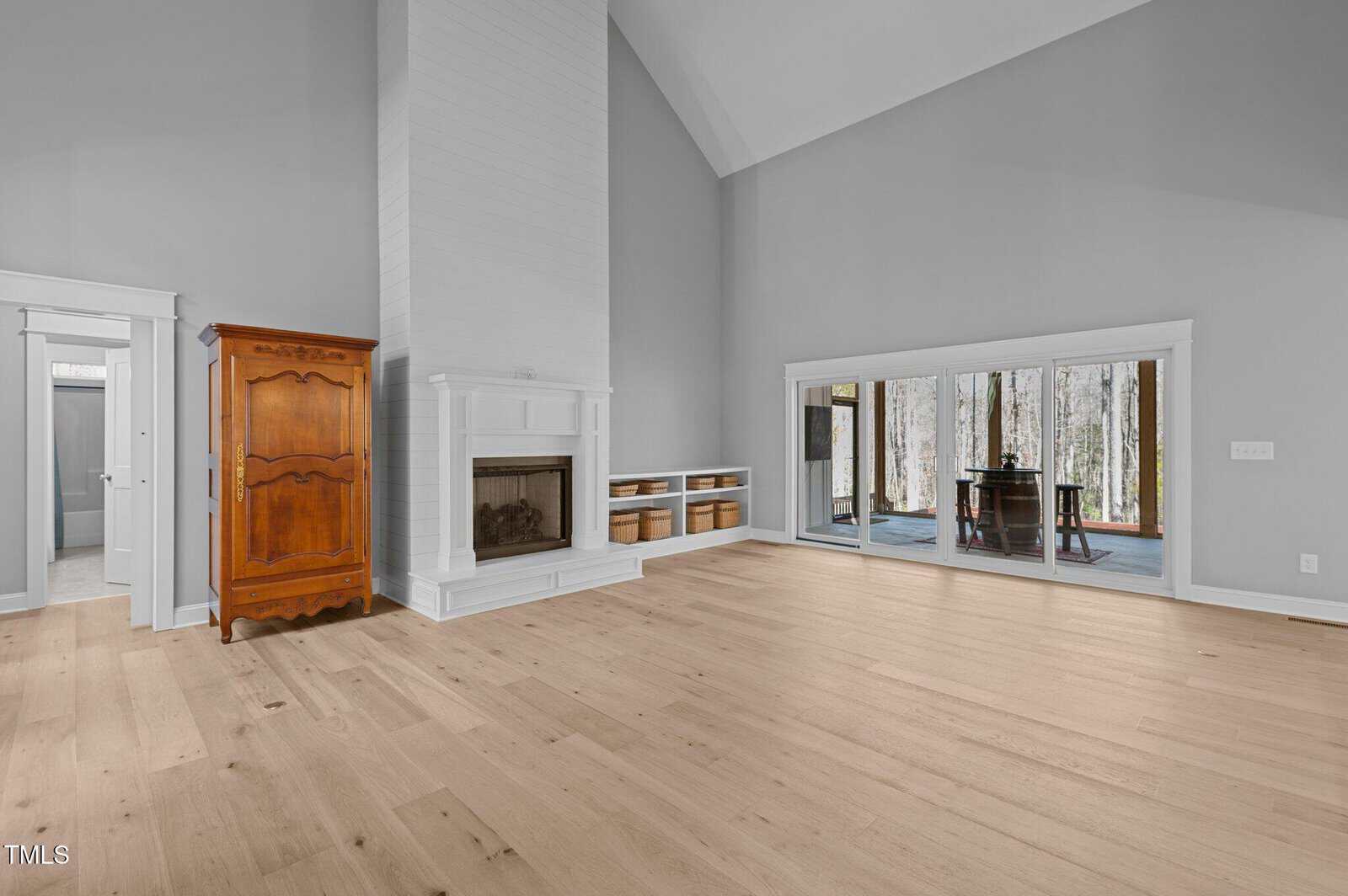 Spacious living room with light wood floors, gray walls, a tall white fireplace, wooden armoire, built-in shelves with baskets, and large sliding glass doors leading to an enclosed porch with a table and chairs.