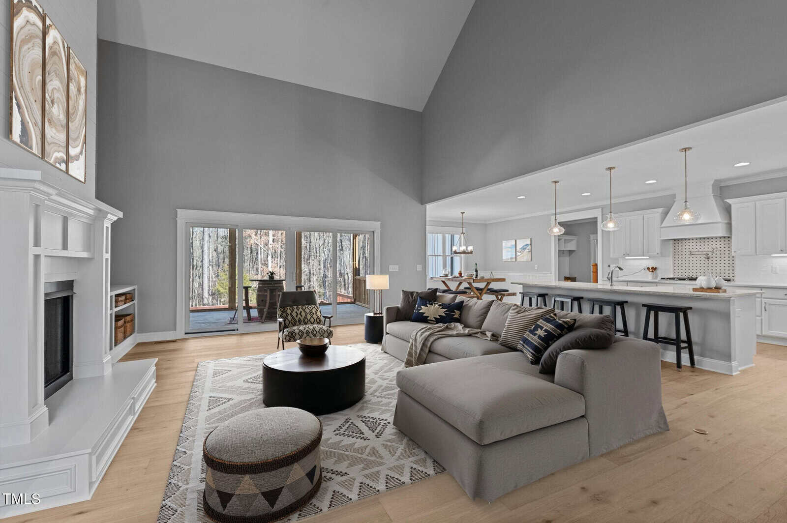 Spacious living room with a gray sectional sofa, patterned rug, round coffee table, and fireplace. Large windows and glass doors open to a deck. Adjacent modern kitchen with pendant lights and a long island.