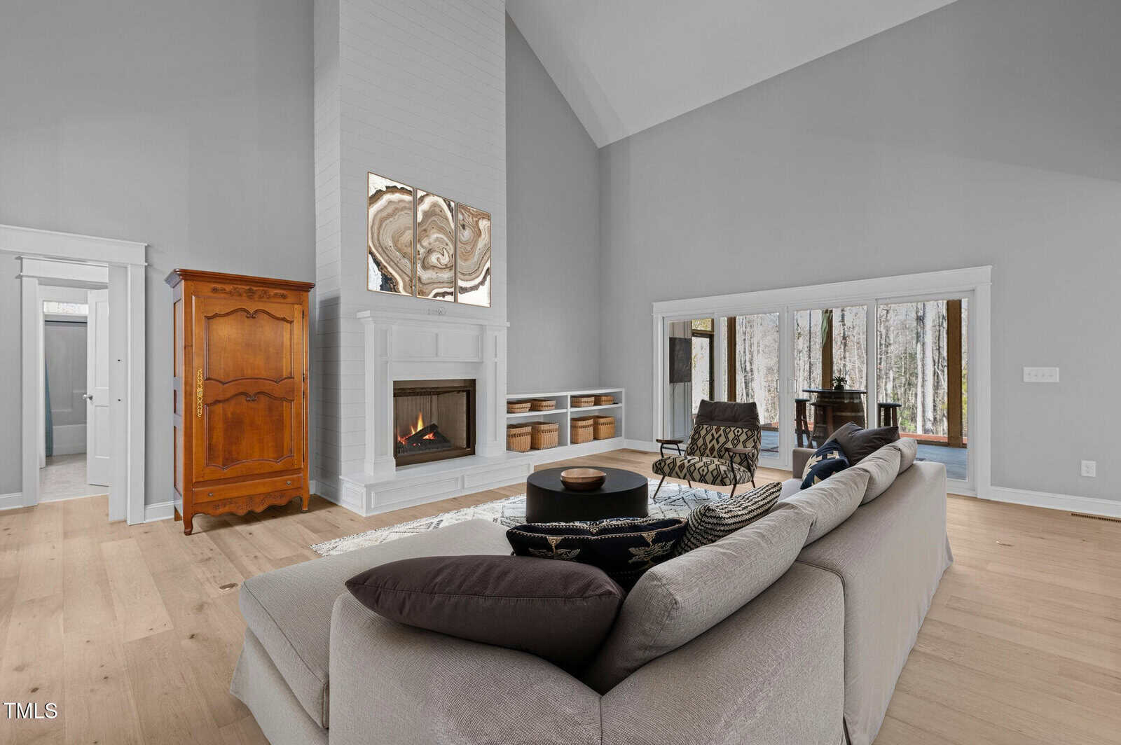 A spacious living room with light wood floors, a white sectional sofa, a modern fireplace, built-in shelves, a wooden armoire, and large windows overlooking a wooded outdoor area.