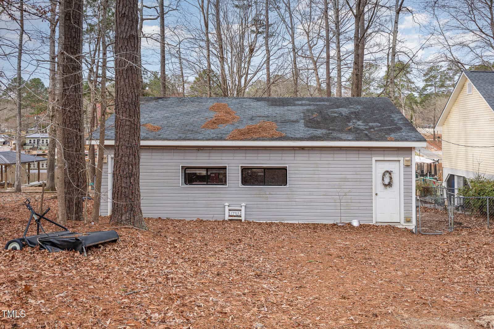 A detached gray garage with three windows, a white door with a wreath, and pine needles scattered on the roof. Fallen leaves and a tipped-over wheelbarrow are visible in the yard. Tall trees surround the area.