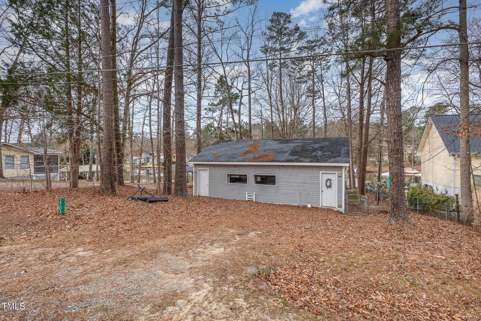 A small gray garage or shed with two windows and a door sits among tall trees. The roof has leaves on it, and the yard is covered in brown pine needles. Other houses are visible in the background.