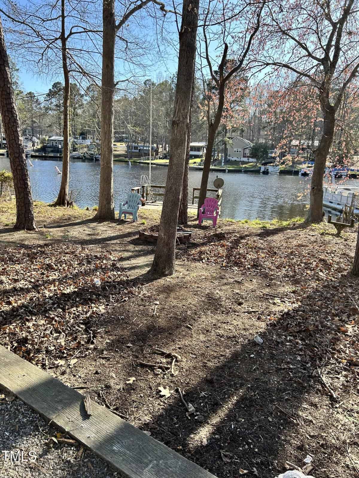 A backyard with several leafless trees, children's chairs, and play equipment overlooks a calm lake with docks and houses visible on the opposite shore, under a clear blue sky.