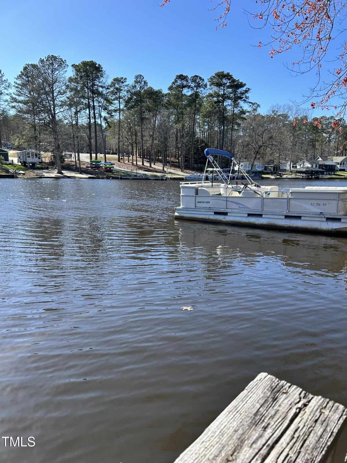 A pontoon boat is docked on a calm lake with trees and several houses visible along the opposite shoreline under a clear blue sky. A wooden pier is in the foreground.
