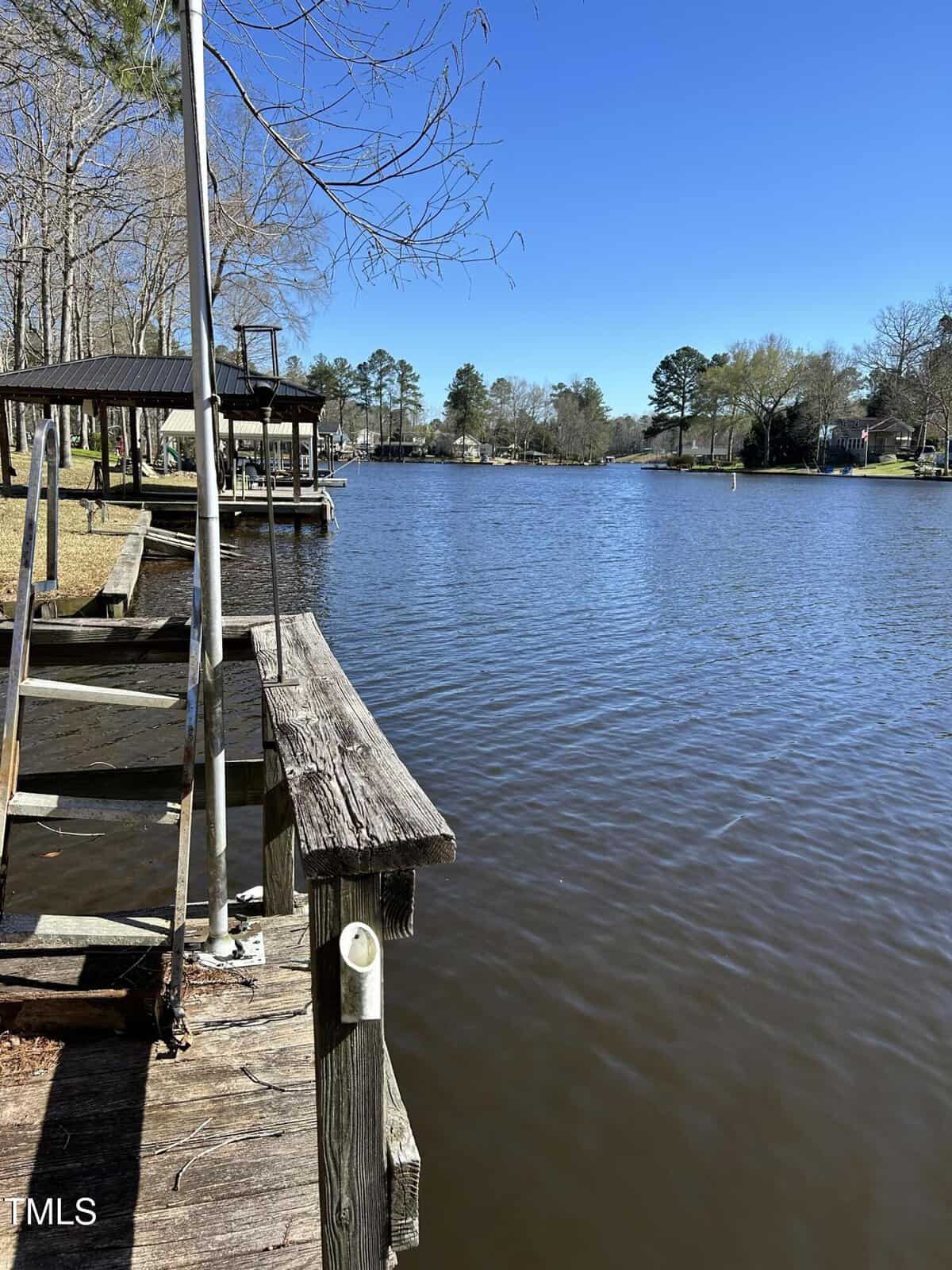 A wooden dock extends over a calm, brownish river with houses and trees lining the opposite bank under a clear blue sky. Some docks and boats are visible along the shoreline.