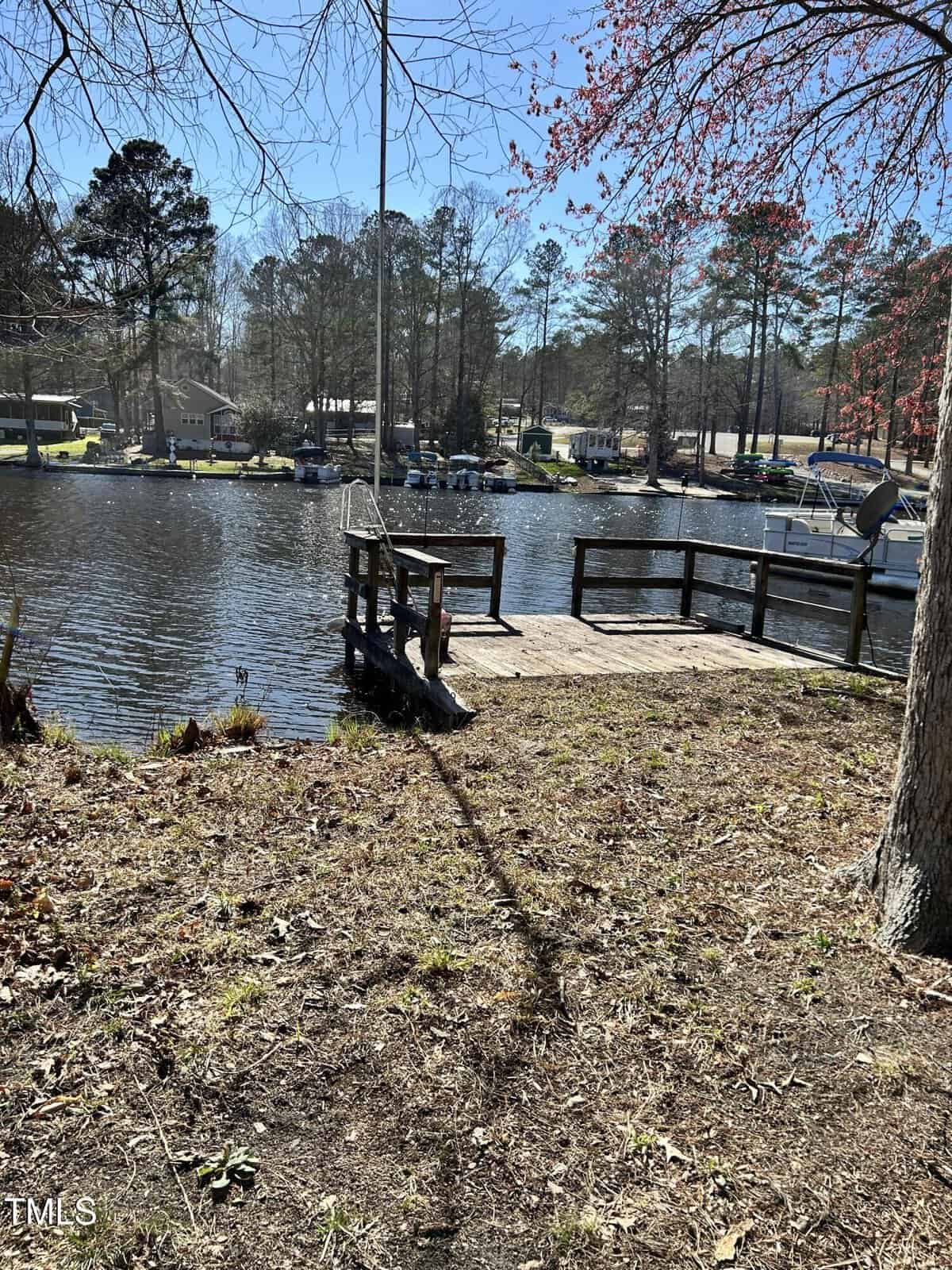 A wooden dock extends over a calm lake with houses and boats visible on the opposite shore. Trees with sparse foliage surround the area under a clear blue sky.