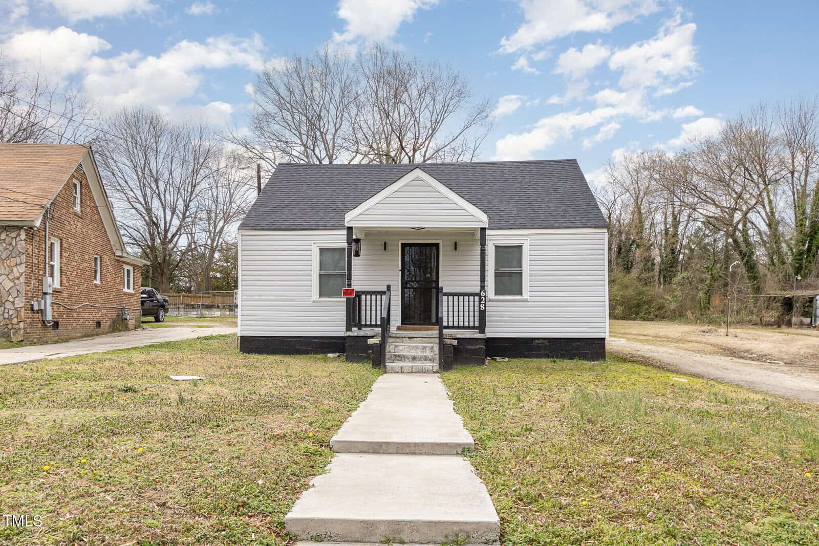 A small, single-story white house with a black roof and front door at 628 East Avenue is shown. It has a porch with steps leading to a concrete walkway. The Henderson home is flanked by trees, with a brick house visible on the left. The sky is partly cloudy.