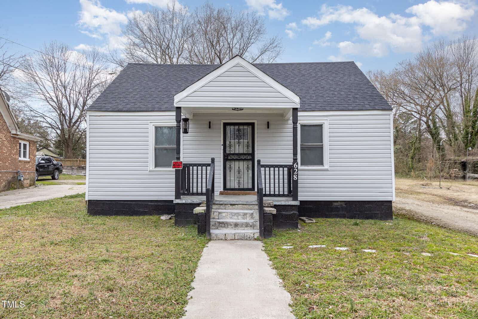 A small, single-story white house with a black roof and trim. It features a porch with steps, black railings, and a black front door with a decorative screen. A paved walkway leads to the entrance, and the yard is grassy with bare trees in the background.