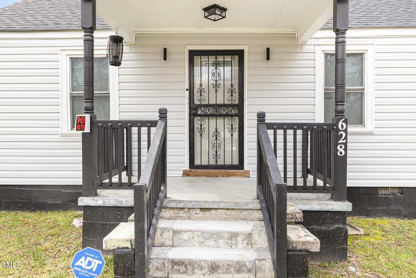 The image shows the front entrance of a house with a small porch. It has a white exterior, black railings, and steps leading up to a black metal door with decorative elements. There is a security sign on the left and the number 628 on the right column.