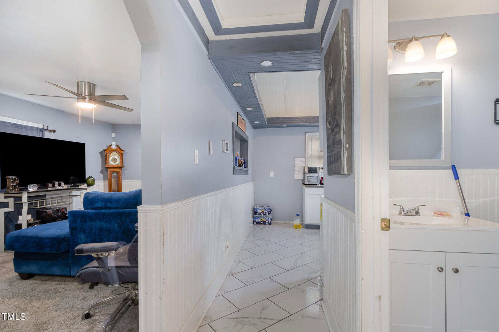 A hallway with light gray walls leads to a living room and a bathroom. The living room has a blue sofa, a grandfather clock, and a flat-screen TV. The bathroom features a sink with a mirror and a cleaning mop on the floor.
