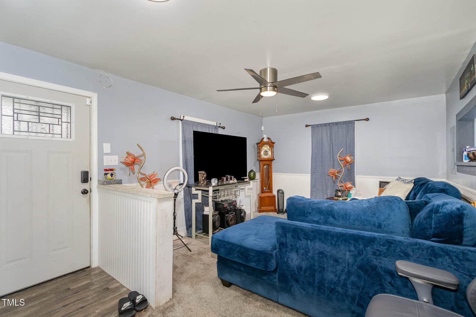 Living room with blue walls and a large blue sectional sofa. A grandfather clock is near the TV, and a ceiling fan with lights hangs overhead. A white door is on the left, and decorative items are placed around the room.