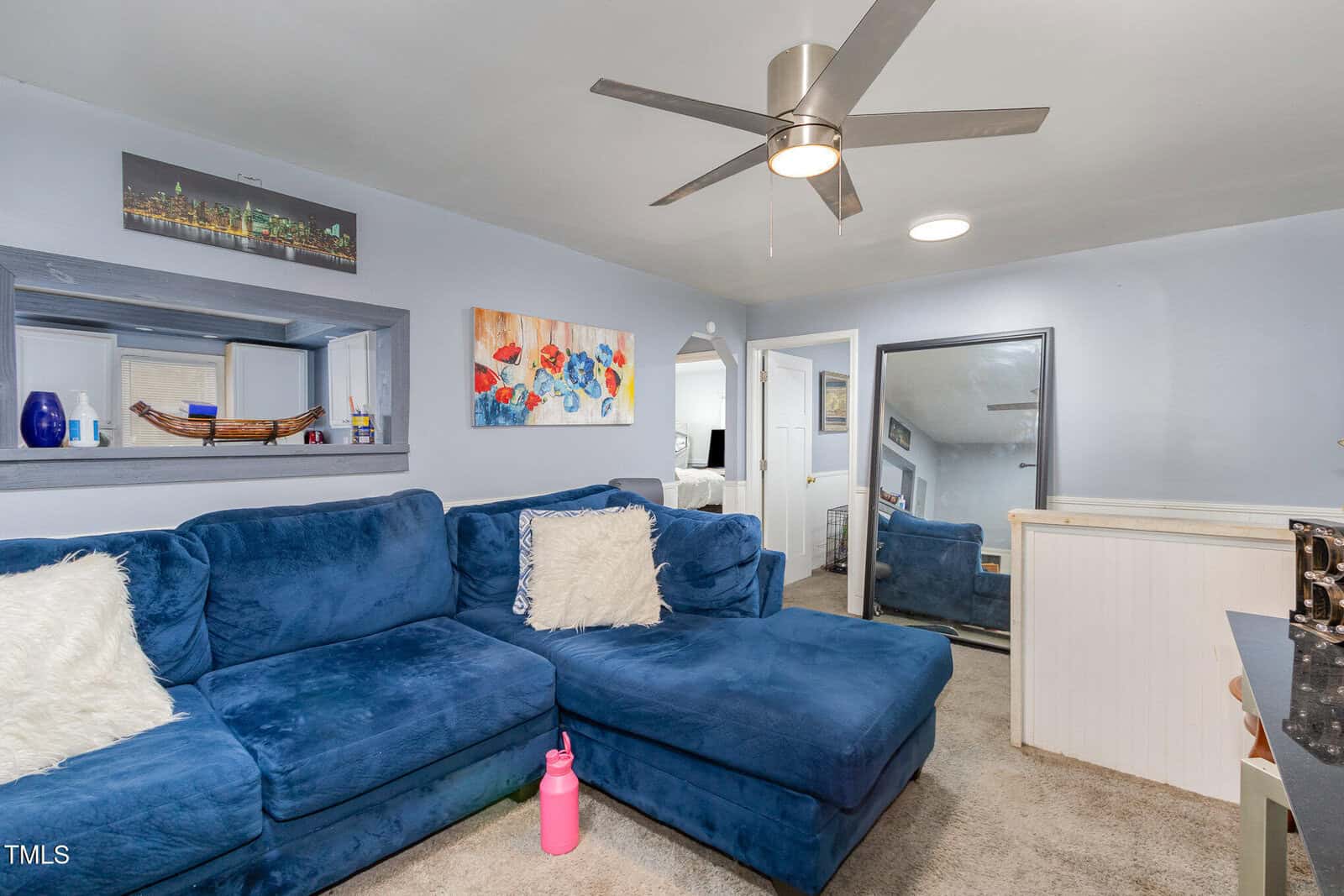 A living room with a large blue sectional sofa and two fluffy white pillows. There are colorful paintings on the light blue walls. A pink water bottle is on the carpet, and there's a ceiling fan above. A mirror leans against one wall.