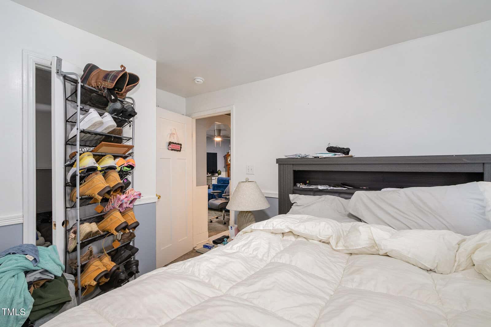 A bedroom with a white comforter on the bed. Shoes are stored on a rack attached to the wall. Clothes are scattered on the floor. There's a lamp on a nightstand, and a doorway opens to another room with visible blue chairs.