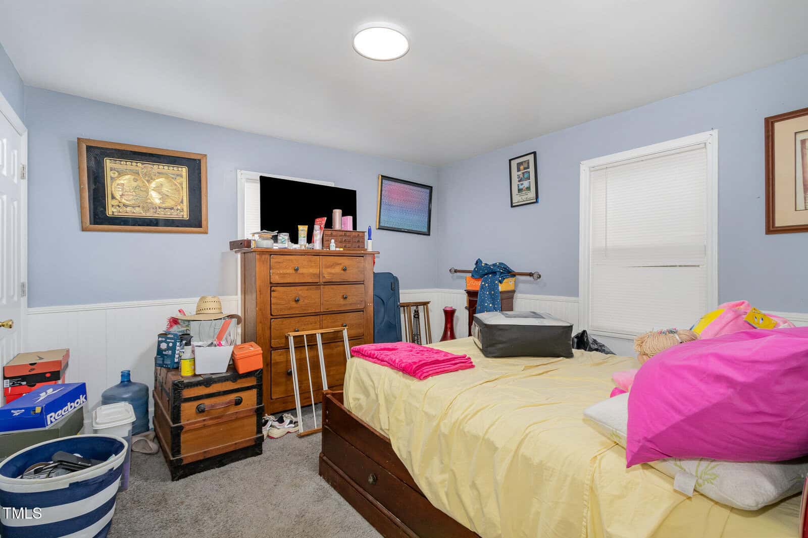 A small bedroom with light blue walls, a bed with yellow sheets and pink pillows, a wooden dresser with various items on top, a window with closed blinds, and miscellaneous items and boxes around the room.