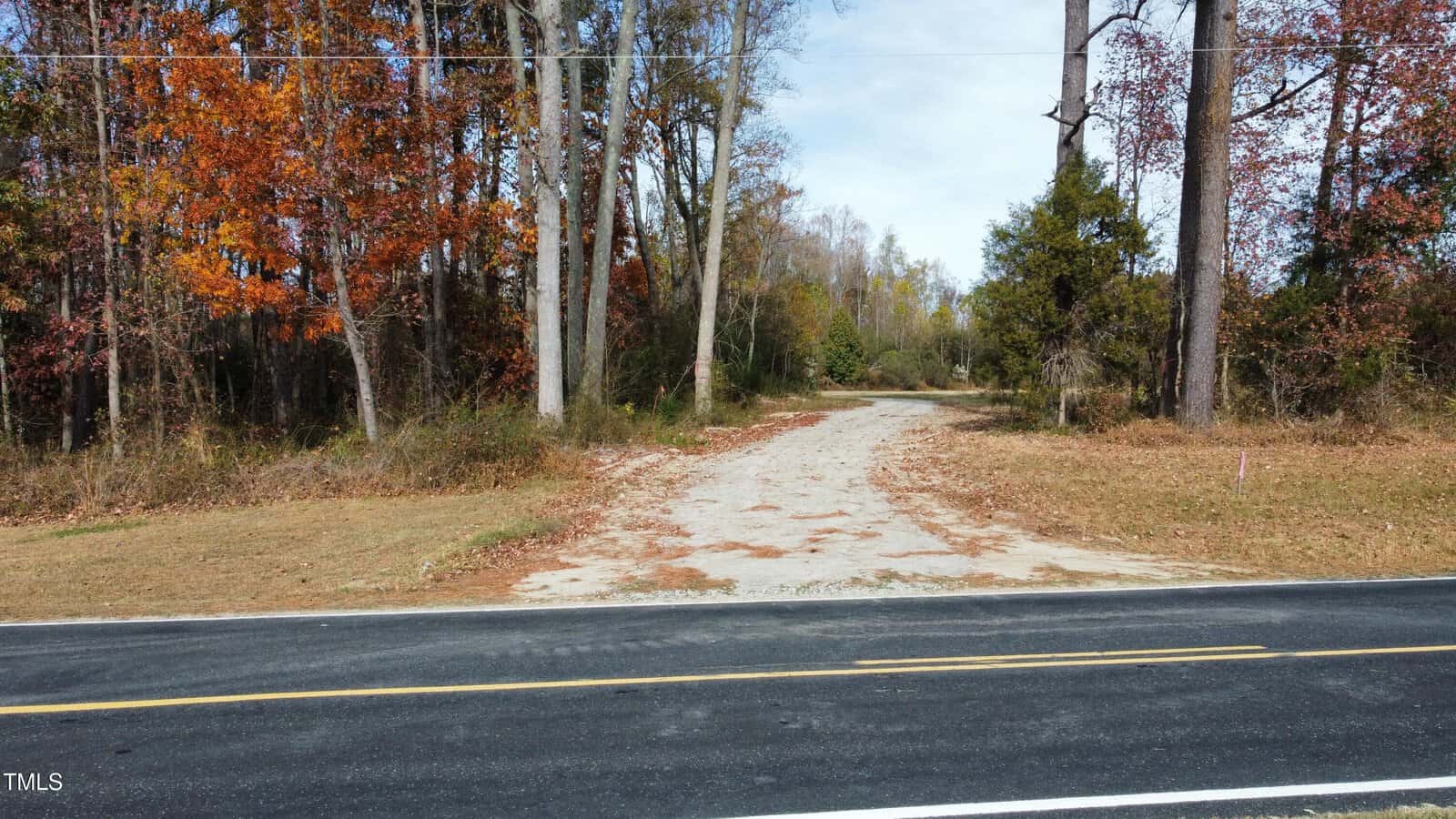 A paved road runs horizontally in the foreground at 0 Duke Memorial Road, Louisburg. Behind it, a gravel driveway leads into a wooded area with trees displaying autumn colors. The sky above is partly cloudy.